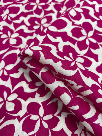 A close-up image of a Super Cheap Fabrics Designer Rayon - Fuchsia - 140cm with a vivid pattern. The design features magenta flowers and white leaves, capturing the vibrant prints beautifully. The fabric appears slightly folded, showcasing its texture and the intricate arrangement of the floral pattern, making it a versatile choice.