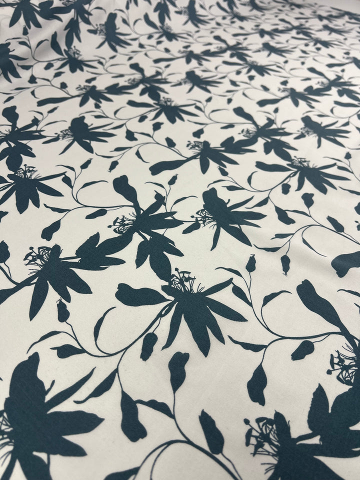 A versatile choice, the Designer Rayon - Smoke Blue - 140cm from Super Cheap Fabrics features a repeating pattern of dark blue, stylized floral silhouettes. The intricate design includes various sizes of flowers, stems, and buds, creating an elegant, nature-inspired motif. The fabric is laid out flat, showcasing the entire pattern.