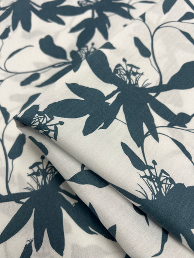 A close-up image of Super Cheap Fabrics' Designer Rayon - Smoke Blue - 140cm with a white background and a dark teal floral pattern. The fabric is slightly folded, showcasing the seamless repetition of the flower design with intricate details in the petals and stems, making it a versatile choice for vibrant prints.