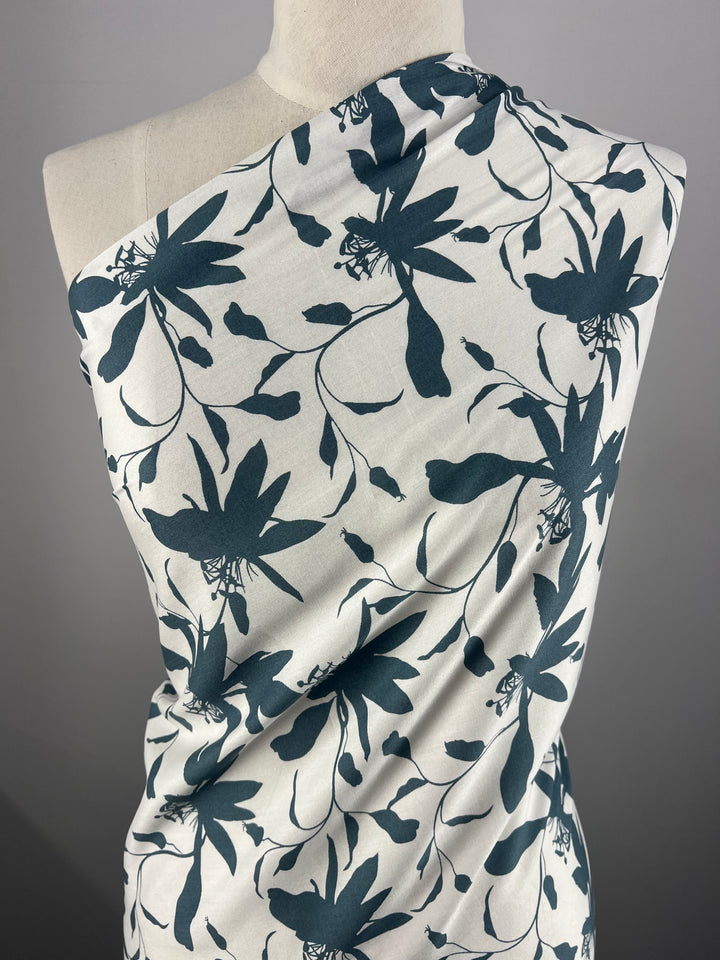 A fabric draped over a mannequin features a white background with vibrant teal botanical prints. The leaves and flower-like shapes create a modern and stylish design. This smooth, lightweight Designer Rayon - Smoke Blue - 140cm from Super Cheap Fabrics is a versatile choice, ideally suited for garments or decor.