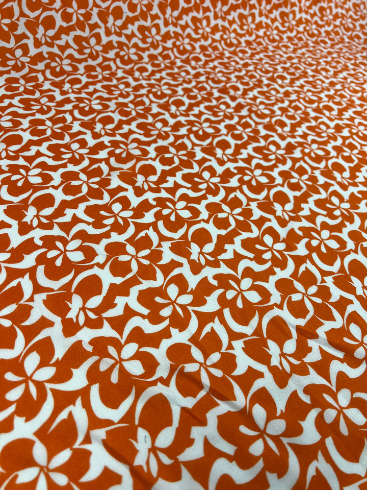 A close-up view of a Super Cheap Fabrics Designer Rayon - Orange Tiger - 140cm with a vibrant orange and white floral pattern. The versatile fabric features stylized flowers closely packed together, creating a textured and visually striking surface.