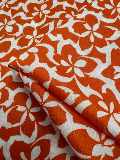 A close-up of Super Cheap Fabrics' Designer Rayon - Orange Tiger - 140cm featuring an orange and white floral pattern. The design consists of stylized flowers with abstract petals in a bold, vivid orange color against a white background. The fabric appears folded, showcasing the repetitive pattern in these vibrant prints.