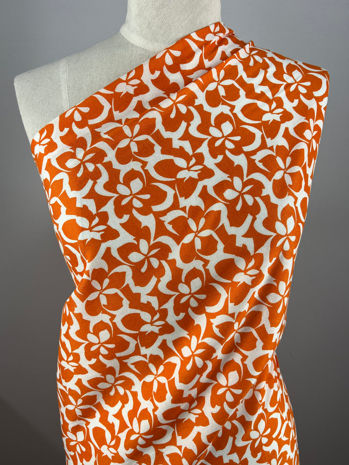 A mannequin is draped with Super Cheap Fabrics' Designer Rayon - Orange Tiger - 140cm featuring a bold, orange floral pattern on a white background. The large, stylized flowers create an eye-catching design, and the versatile fabric is arranged to cover the mannequin like a one-shoulder dress.