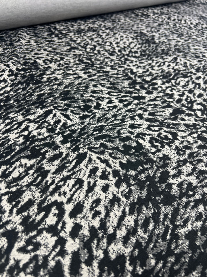 A close-up of a medium weight fabric roll featuring a black and white abstract pattern. The design resembles irregular, dense clusters of brush strokes, creating a textured, speckled effect across the surface. The product shown is Super Cheap Fabrics' Textured Knit - Snow Leopard - 145cm.