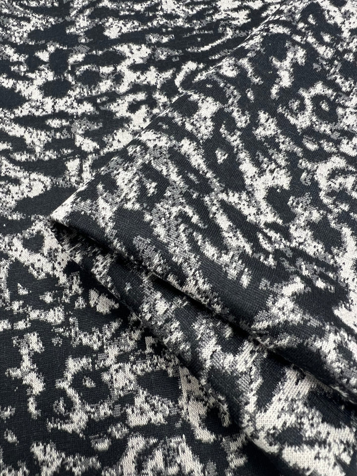 A close-up view of a medium weight fabric with a black and white abstract pattern. The Textured Knit - Snow Leopard - 145cm from Super Cheap Fabrics appears to have a soft, flexible material with intricate detailing in the design. The image focuses on the surface, showcasing the intricacy and contrast of the pattern.