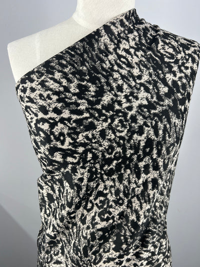 A close-up of a mannequin draped with the Textured Knit - Snow Leopard - 145cm by Super Cheap Fabrics, featuring a black and white abstract pattern resembling animal print. The textured knit fabric's intricate design and generous stretch are prominently displayed against a plain background.