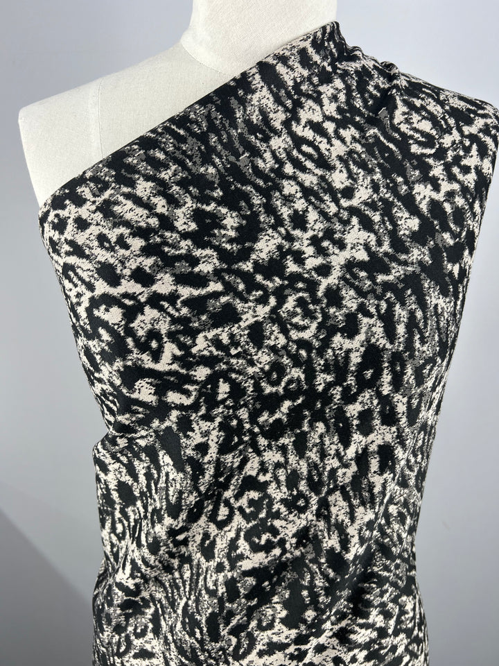 A close-up of a mannequin draped with the Textured Knit - Snow Leopard - 145cm by Super Cheap Fabrics, featuring a black and white abstract pattern resembling animal print. The textured knit fabric's intricate design and generous stretch are prominently displayed against a plain background.