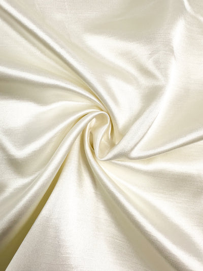 A close-up of the Shantung Satin - Pristine fabric from Super Cheap Fabrics, showcasing its smooth, light-colored surface adorned with a swirl pattern at the center. Made from 100% polyester, this material has a shiny, glossy texture that reflects light beautifully, evoking a sense of luxury. The lightweight nature of this satin swirl suggests a soft and flowing movement. The fabric is 150cm wide.