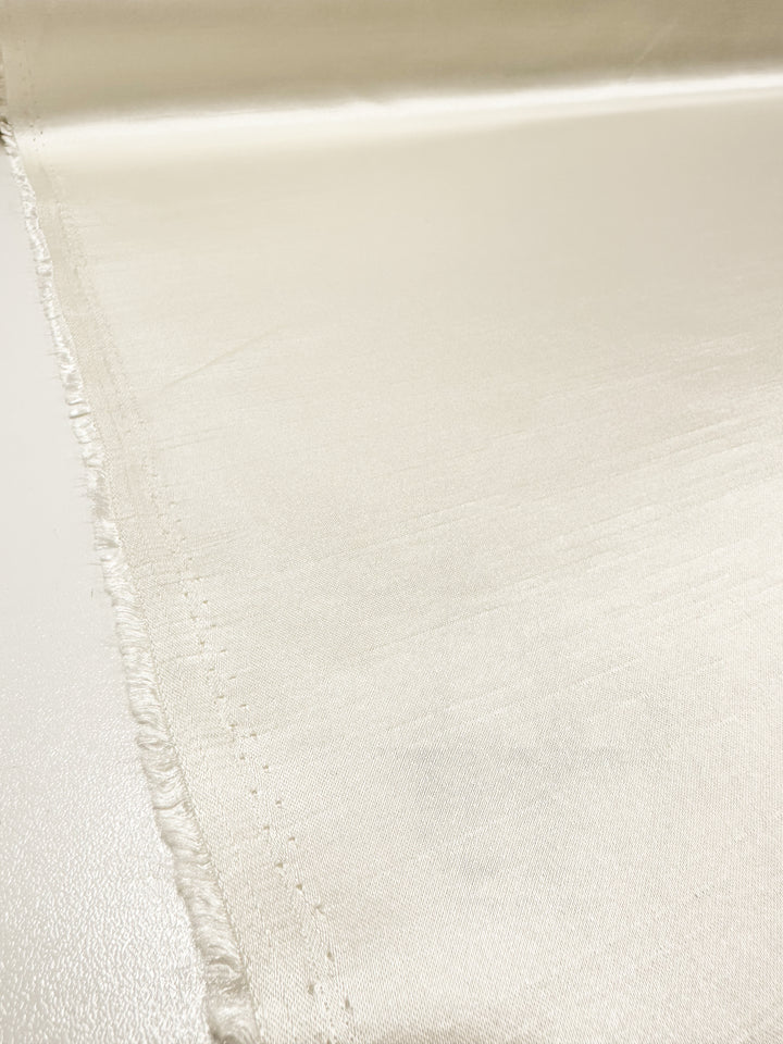 A close-up view of a piece of light cream-colored, lightweight Shantung Satin - Pristine - 150cm fabric from Super Cheap Fabrics laid out flat. The 100% polyester material has a smooth, shiny surface with a soft texture and slightly frayed edges on one side.