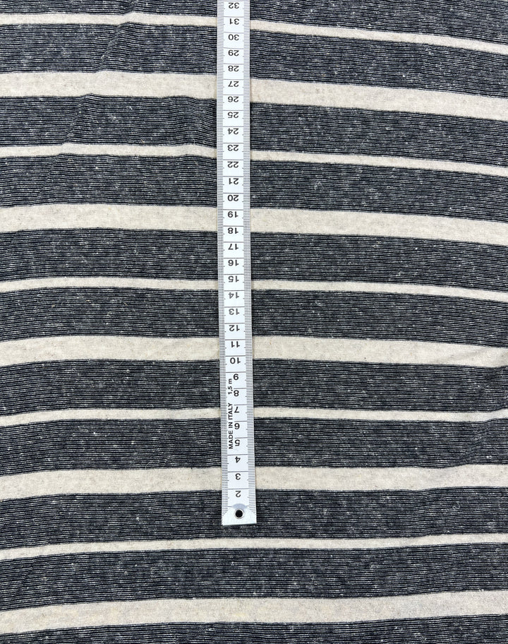 A measuring tape placed vertically on a dark fabric with white horizontal stripes. The tape shows measurements in inches, ranging from 1 to 27 inches. The Linen Jersey - Sand Stripe - 150cm by Super Cheap Fabrics has a woven texture and the stripes are evenly spaced.
