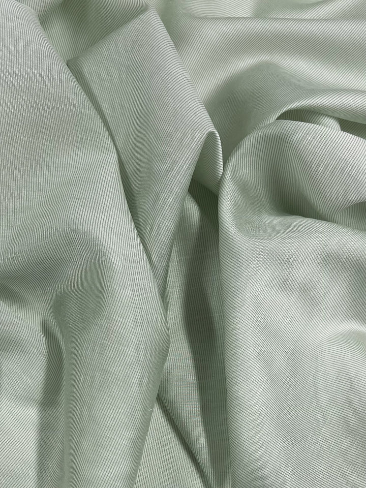 A close-up image of light green Designer Silk - Prodigy - 158cm by Super Cheap Fabrics with fine, subtle stripes. The material is softly draped, creating gentle folds and shadows, which add texture and depth to the lightweight fabric's surface. Ideal for crafting elegant dresses.
