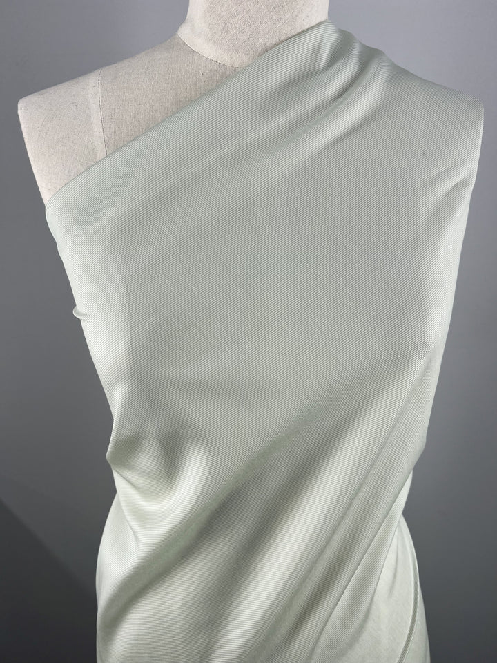 A mannequin draped with a smooth, light green, one-shoulder Designer Silk - Prodigy - 158cm fabric from Super Cheap Fabrics. The lightweight material appears soft and has a slight sheen, showcasing subtle folds and texture. The background is plain and gray, emphasizing the simplicity and elegance of the draped fabric.