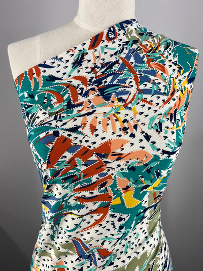 A one-shoulder dress displayed on a mannequin features a vibrant, multicolored abstract pattern with bold reds, oranges, greens, blues, and yellows on a white background. Made from Super Cheap Fabrics' medium-weight Printed Lycra - Dotted Forrest - 150cm fabric, the dress has a form-fitting design and an artistic, dynamic print.
