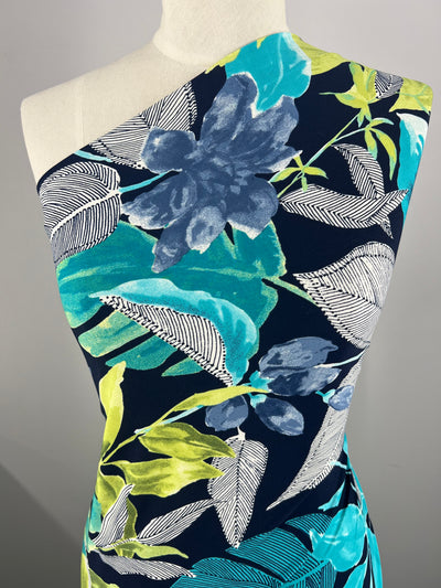 A dress on a mannequin featuring a bold tropical print with large leaves and flowers in shades of blue, green, yellow, and white on a dark background. The medium-weight fabric is the Super Cheap Fabrics Printed Lycra - Flowy Foliage - 150cm, adding stretch to its asymmetrical one-shoulder design.