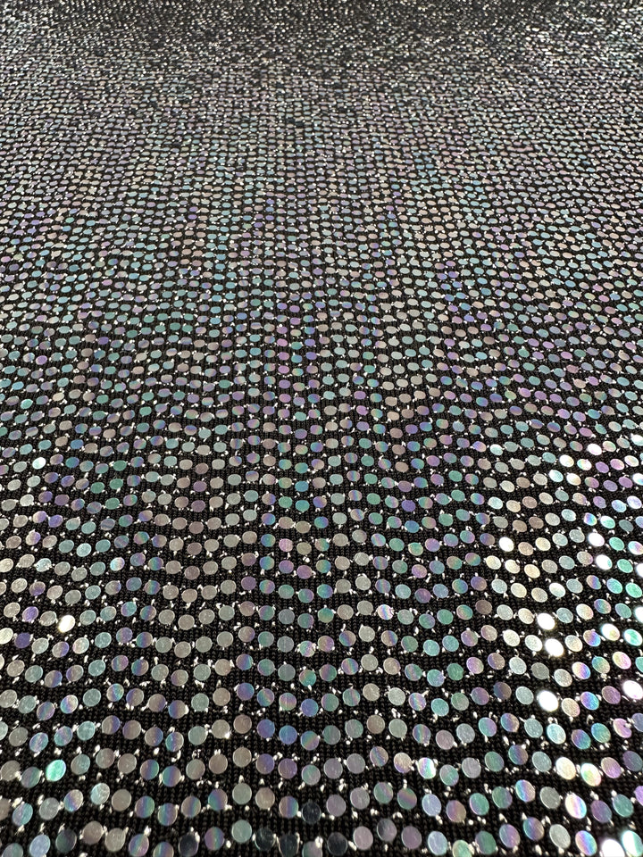 A close-up view of the Designer Sequins - Disco - 145cm fabric from Super Cheap Fabrics, showcasing a dense pattern of small, circular sequins on black and multi fabric. This 100% polyester material has a reflective, iridescent quality that displays a spectrum of colors including green, purple, and silver which shift with the angle of light.