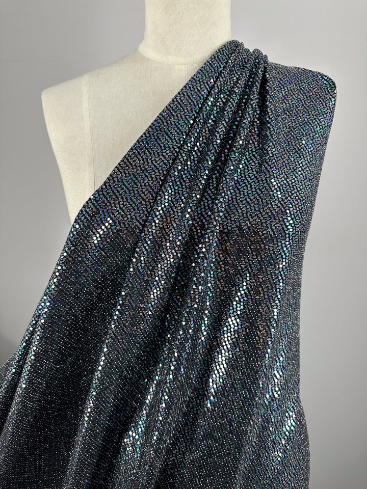 A mannequin draped in Super Cheap Fabrics' Designer Sequins - Disco, a shimmering, medium to heavy weight fabric that reflects light with an iridescent effect. The black and multi-colored, 100% polyester material features a scale-like pattern that covers the mannequin from one shoulder across the torso. The fabric's sparkle is highlighted by a neutral gray background.