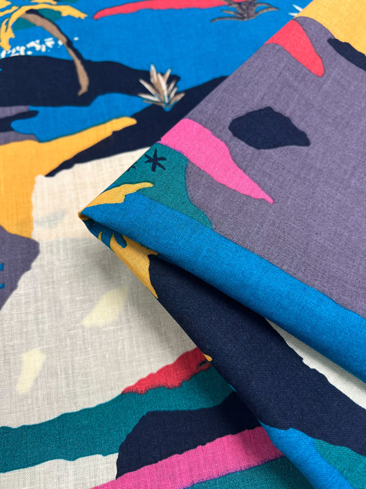 A close-up image shows two pieces of colorful, lightweight fabric layered over each other. The Linen Cotton - Palm Tree - 135cm by Super Cheap Fabrics features abstract, tropical patterns in vibrant colors, including blue, pink, yellow, and green. Palm trees and other shapes are also visible in the design.