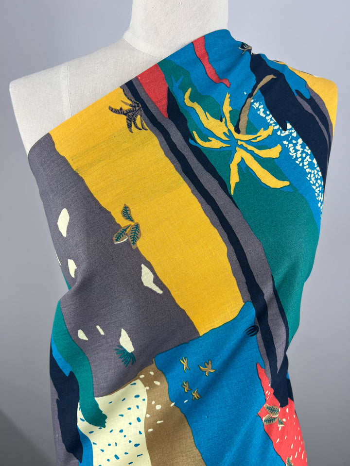 A close-up of a multicolored textile draped over a mannequin. The light weight fabric features an abstract design with a mix of yellow, green, blue, red, and gray panels, along with depictions of leaves and small birds. This Super Cheap Fabrics' Linen Cotton - Palm Tree - 135cm piece rests gracefully against the plain gray background.