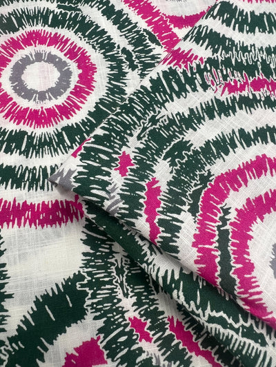 A close-up of Linen Cotton - Fuzz - 147cm by Super Cheap Fabrics featuring a bold, colorful pattern with concentric circles. The circles are in shades of magenta, gray, and dark green with a white background, creating a vibrant and eye-catching design. The breathable fabric appears to be folded, showing multiple layers.