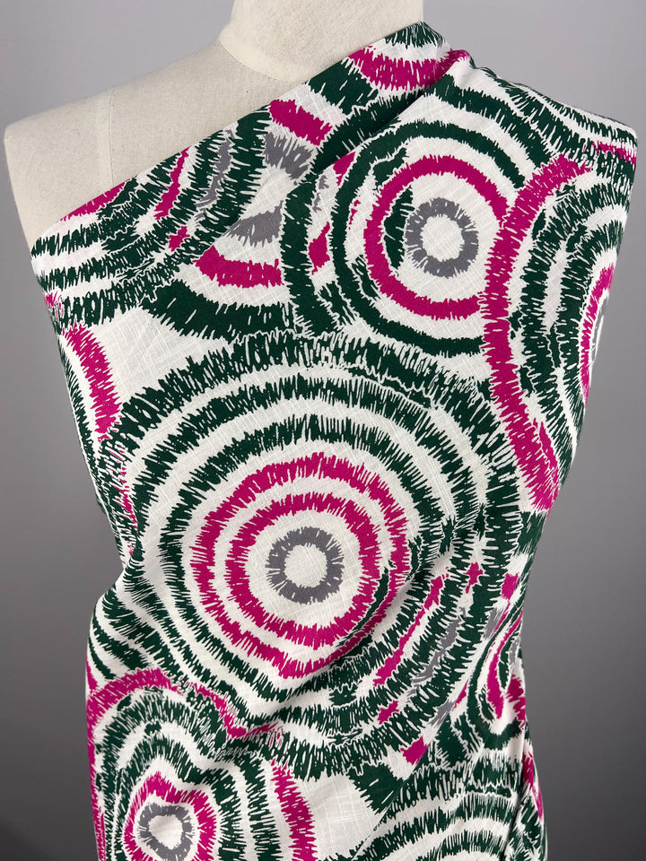 A mannequin is draped in durable fabric featuring a pattern of concentric circles. The circles are primarily dark green, pink, and grey, creating a vibrant and dynamic visual effect. The Linen Cotton - Fuzz - 147cm by Super Cheap Fabrics is wrapped diagonally across the mannequin's shoulder and torso.