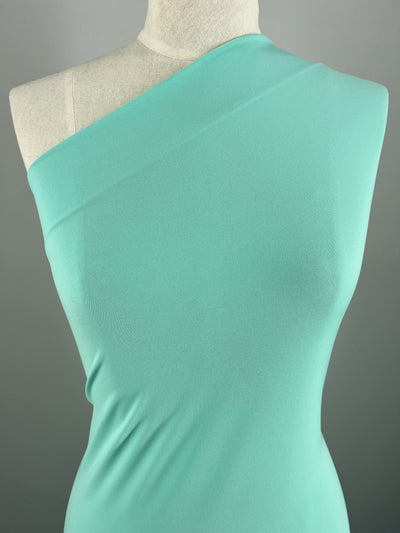 A mannequin dressed in an Ice Blue, one-shoulder dress made from Super Cheap Fabrics' ITY Knit - Ice Blue - 150cm fabric. The medium weight fabric drapes smoothly, showcasing the elegant and modern design of the dress. The background is a plain, muted gray, drawing attention to the vibrant color and sleek details of the garment.