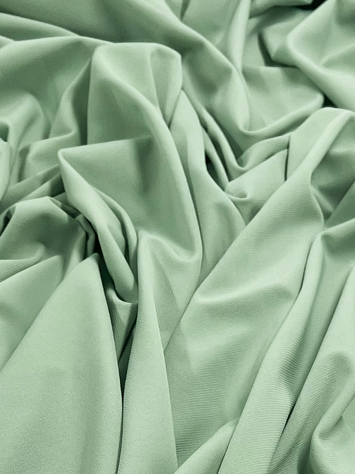 A close-up of soft, crumpled mint-green interlock twist yarn fabric from Super Cheap Fabrics with gentle folds and wrinkles, creating a textured appearance and showcasing the smooth, silky 95% polyester material of the ITY Knit - Sage - 150cm.