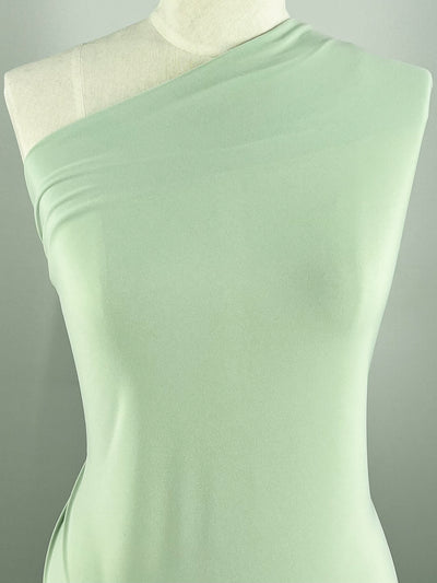 A mannequin is draped in light green, medium weight fabric. The smooth 95% polyester material, ITY Knit - Sage - 150cm by Super Cheap Fabrics, is wrapped to create an asymmetrical, one-shoulder look, highlighting the mannequin's upper body. The background is neutral, ensuring the elegant fabric stands out beautifully.