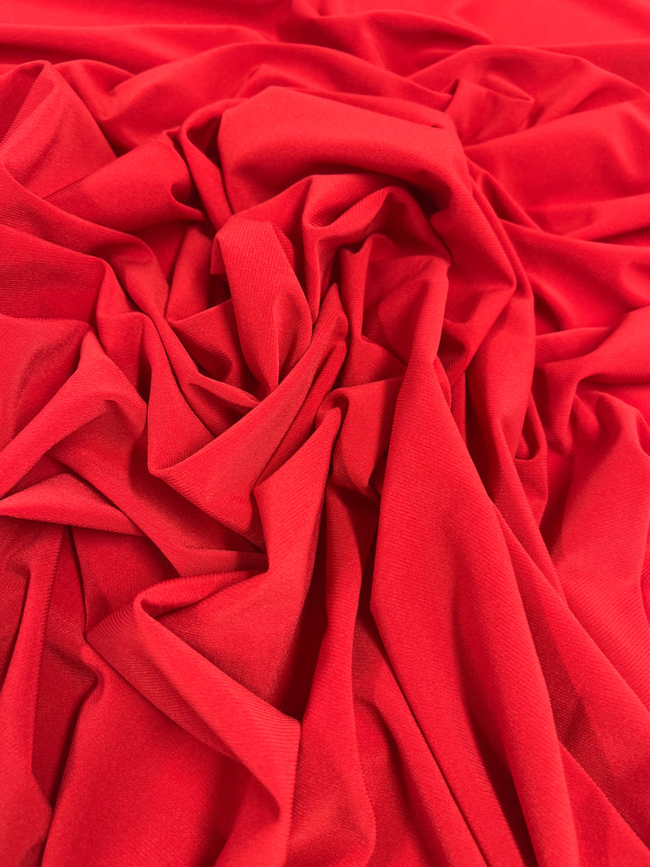 A close-up of a vibrant red ITY Knit - Red - 150cm fabric with a smooth, slightly shiny texture from Super Cheap Fabrics. The medium weight fabric is laid out in a series of intricate folds and creases, creating a dynamic array of shadows and highlights.