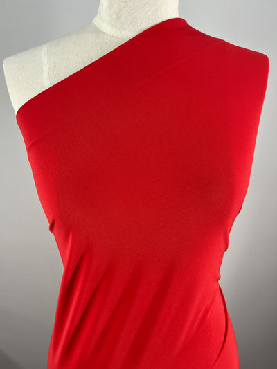 A mannequin is draped with ITY Knit - Red - 150cm from Super Cheap Fabrics, displaying the elegant and smooth texture of the material. The background is plain and grey, focusing attention on the vibrant red fabric and its graceful draping.