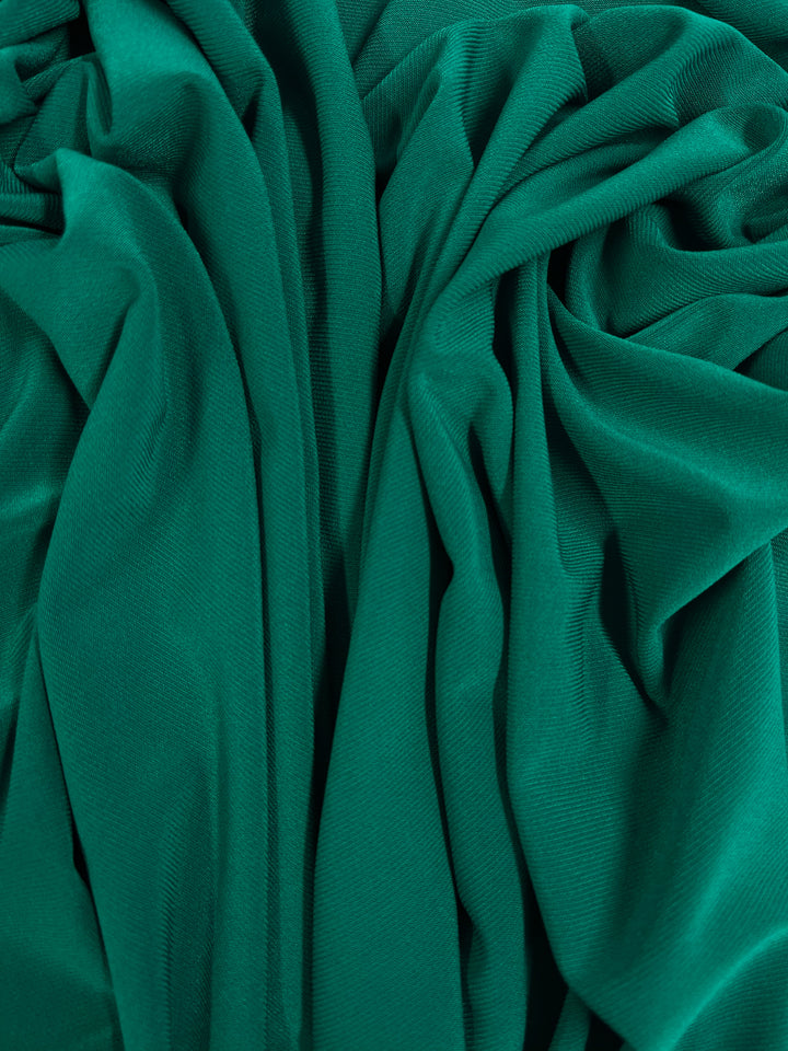 A close-up photo of vibrant, dark teal fabric with intricate, loose folds and pleats, creating a textured and dynamic appearance. The medium weight ITY Knit - Dark Teal - 150cm from Super Cheap Fabrics appears soft and silky, capturing light and shadow to accentuate the depth of the folds.
