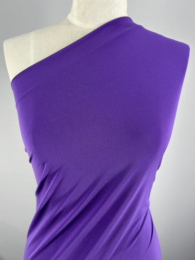 A close-up of a mannequin draped in a vibrant ITY Knit - Pop Purple - 150cm from Super Cheap Fabrics, one-shoulder garment made from a Polyester Spandex blend. The medium weight fabric appears smooth and form-fitting, emphasizing the elegance of the design. The background is plain and light-colored, ensuring the focus remains on the garment.
