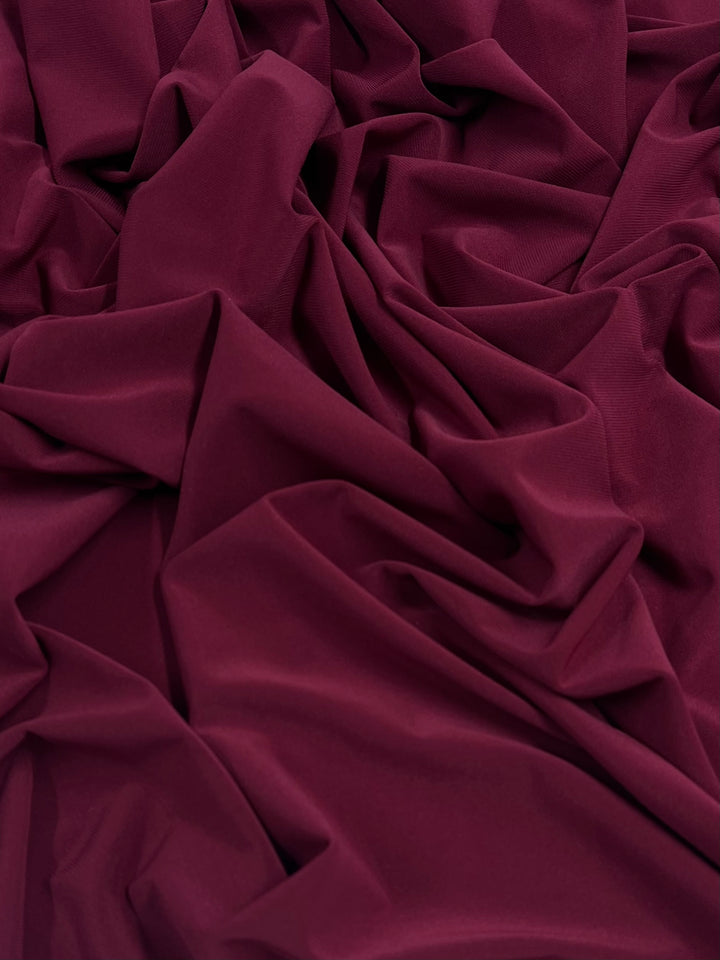 A close-up of a spread of deep merlot-colored fabric. The ITY Knit - Merlot - 150cm by Super Cheap Fabrics has multiple folds and creases, creating a textured appearance. The medium weight material appears soft and smooth to the touch.