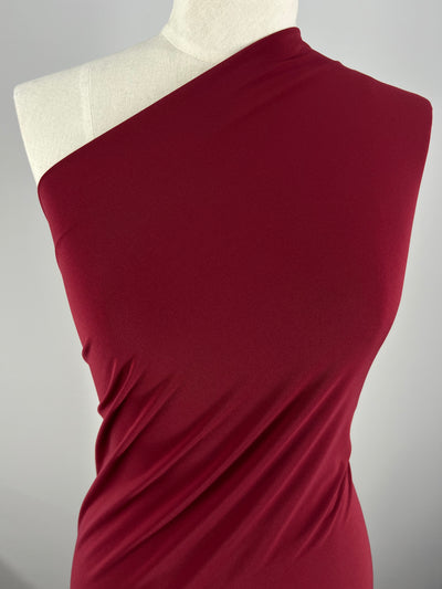 A close-up of a mannequin dressed in a one-shoulder, deep red garment made from medium weight fabric that drapes elegantly. The smooth texture and rich, vibrant color are accentuated by the plain, neutral backdrop, emphasizing the striking hue of tops and dresses crafted from Super Cheap Fabrics' ITY Knit - Merlot - 150cm material.