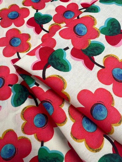 A close-up shot of Printed Cotton - Red Glass Flowers - 150cm by Super Cheap Fabrics featuring a vibrant floral design. The material, made from 100% cotton, showcases large pink and red flowers with blue centers and green leaves on stems, set against a white background. The natural cotton fibers appear soft and folded neatly.