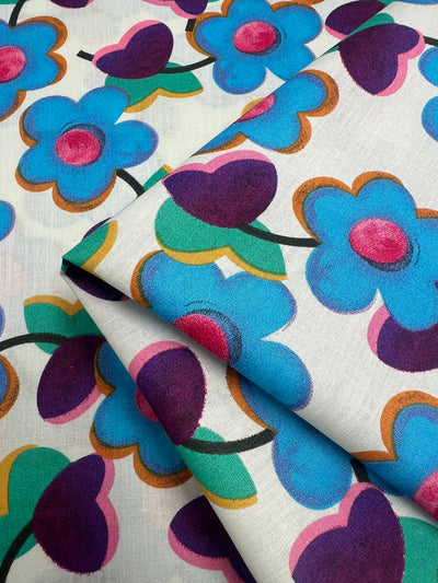 A close-up image of vibrant fabric featuring a floral pattern. The design includes large blue, purple, and pink flowers with green leaves against a white background. Made from natural cotton fibers, the fabric is neatly folded, showcasing the colorful and lively print perfect for multi-use textiles. The product shown is Printed Cotton - Blue Glass Flowers - 150cm by Super Cheap Fabrics.