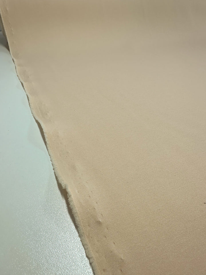 A detailed view of the Scuba Knit - Mother of Pearl - 165cm from Super Cheap Fabrics shows its beige, heavyweight material with a smooth texture spread out on a surface. The fabric has a slightly uneven edge on the left side and appears unfinished or uncut along that side. The underlying surface is made of clean, light-colored material.