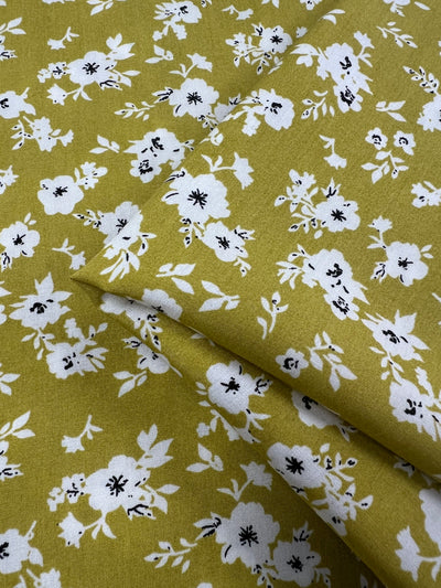 A folded piece of yellow, lightweight fabric adorned with a white floral pattern. The flowers are scattered across the Super Cheap Fabrics' Printed Rayon - Geranium - 145cm, featuring small black accents in their centers. The overall design is light and airy, creating a cheerful and vibrant appearance.