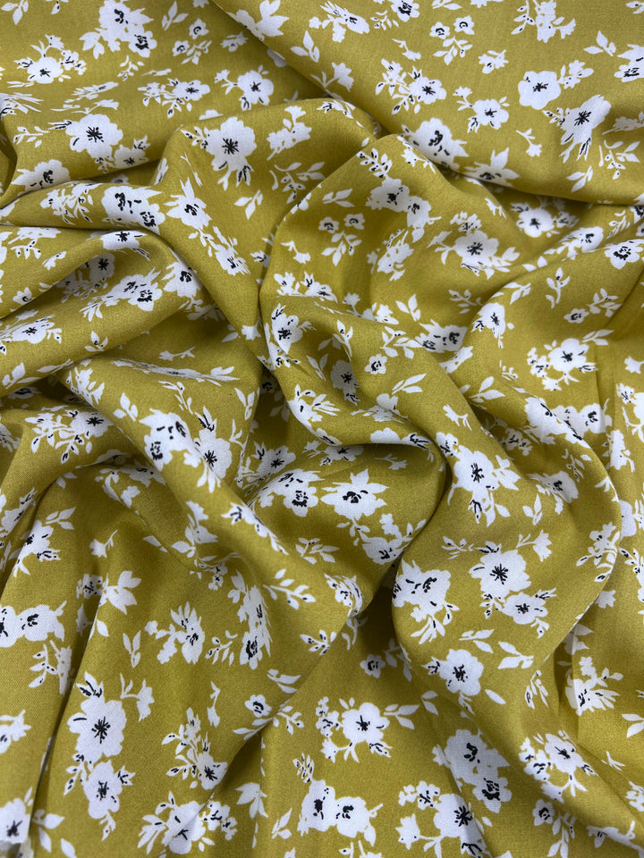 A close-up of crumpled olive green fabric adorned with white floral patterns. The flowers vary in size and have black accents at their centers, creating a contrasting and vibrant design. This lightweight fabric has a soft and flowing texture, characteristic of Super Cheap Fabrics' Printed Rayon - Geranium - 145cm.