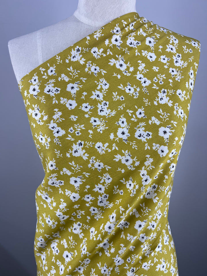 A mannequin draped with Printed Rayon - Geranium - 145cm by Super Cheap Fabrics, adorned with vibrant small white flowers and green leaves, displayed against a gradient blue-gray background. The lightweight fabric is arranged to showcase its texture and design, covering one shoulder of the mannequin.