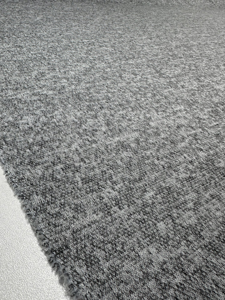 A close-up of Super Cheap Fabrics' Textured Knit - Grey Marle - 155cm shows its gray, textured surface—like medium-weight knit fabric—at the edge where it meets a smooth, lighter-colored floor.