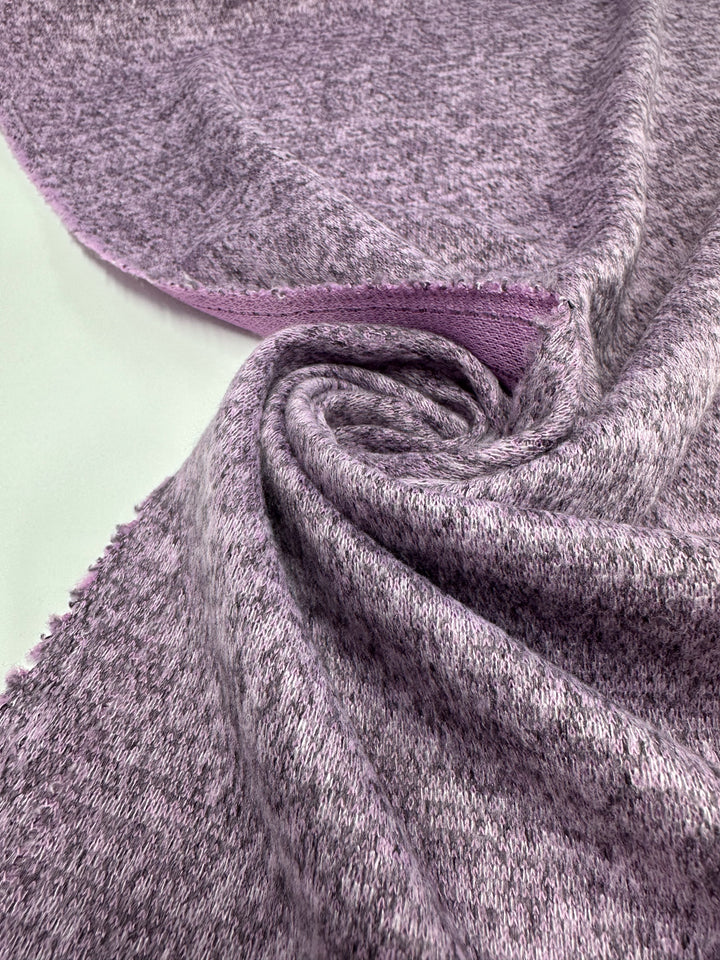 A close-up of Super Cheap Fabrics’ Textured Knit - Orchid Marle - 150cm, medium weight with a soft feel, heathered purple color and smooth reverse, partially twisted and displayed on a white background.