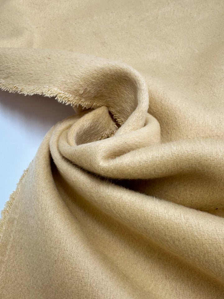 A close-up of Super Cheap Fabrics' Wool Cashmere - Banana - 150cm shows its luxuriously soft, slightly fuzzy beige surface with gentle twists and frayed edges, all set against a white background.