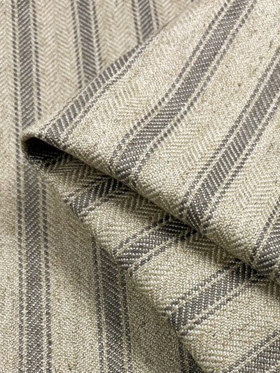 A close-up image of folded *Upholstery Herringbone - Silent Storm - 145cm* fabric by *Super Cheap Fabrics*, showcasing a beige background with dark grey, narrow, parallel stripes. The texture appears woven, with a visible herringbone pattern evident between the stripes for added detail. Part of our affordable range, it's crafted from durable fabrics for lasting quality.