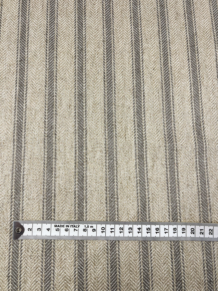 A close-up view of an Upholstery Herringbone - Silent Storm - 145cm fabric with a repeating herringbone pattern in beige and dark gray stripes. A white measuring tape at the bottom shows centimeter markings from 0 to 21, with the text "MADE IN ITALY." This durable fabric from Super Cheap Fabrics is perfect for various applications.