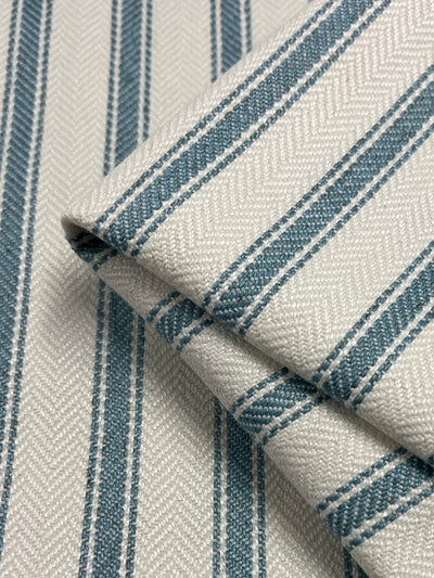 A close-up image of folded Upholstery Herringbone - Glacier Lake - 145cm by Super Cheap Fabrics featuring a pattern of light gray and white stripes against a textured background. The stripes are parallel and evenly spaced, with a subtle herringbone design woven into the durable, stylish fabric.