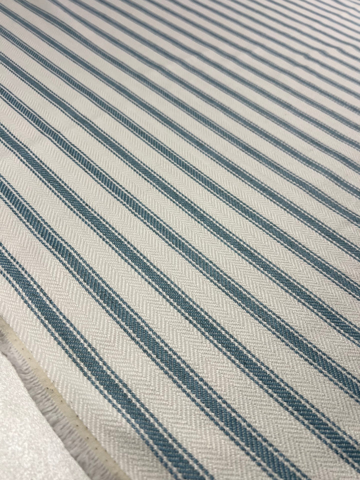 Close-up view of an Upholstery Herringbone - Glacier Lake - 145cm by Super Cheap Fabrics with a pattern of alternating blue and off-white diagonal stripes. The material has a textured weave, and the stripes create a visually dynamic, zigzag effect. The durable stylish fabric appears to be laid out flat.