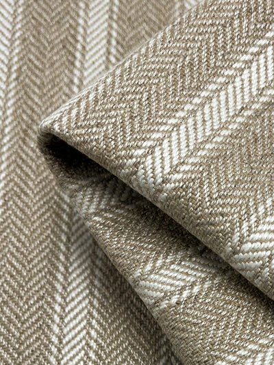 A close-up image of beige and white herringbone patterned Upholstery Herringbone - White & Cream - 145cm by Super Cheap Fabrics. The fabric is folded, showcasing the diagonal zigzag weave design. This durable fabric has a soft and detailed texture, making it an excellent choice within an affordable range.
