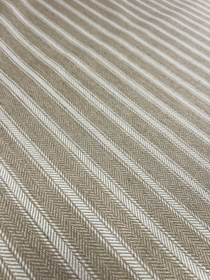 A close-up view of a durable upholstery fabric with a herringbone pattern. The Upholstery Herringbone - White & Cream - 145cm from Super Cheap Fabrics features alternating white and beige diagonal lines, creating a texture that resembles interlocking V-shaped weaving. The focus is on the detailed stitching and pattern consistency in this affordable range.
