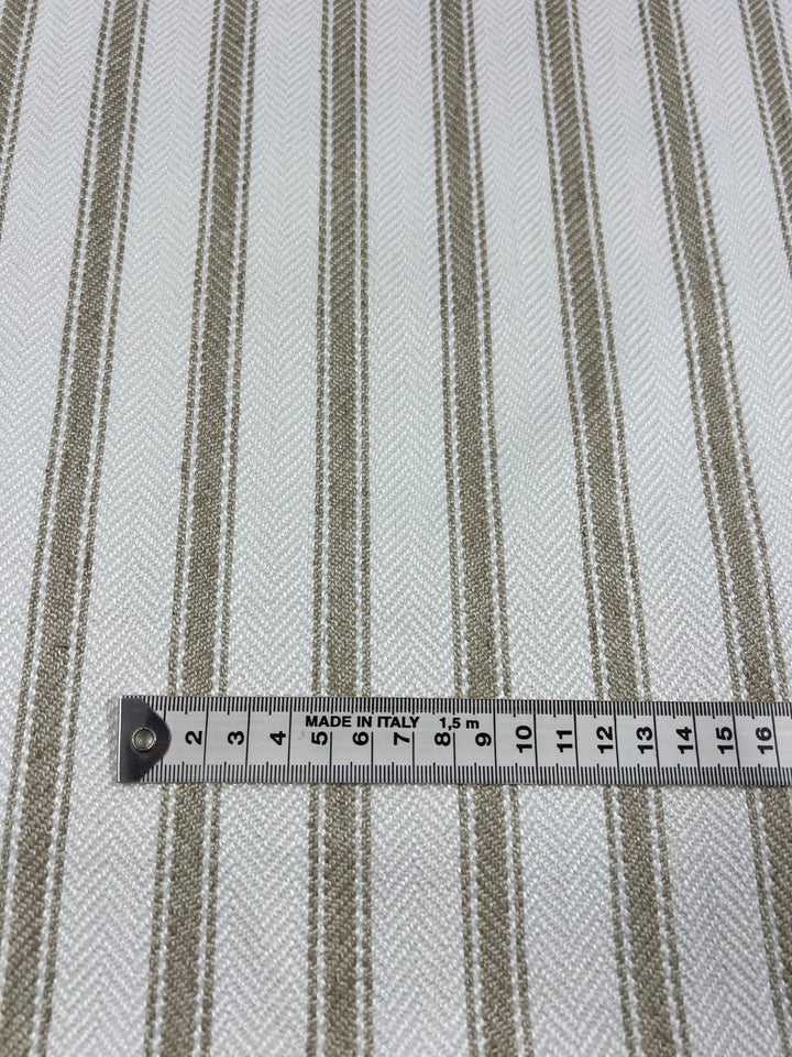 A close-up of Upholstery Herringbone - Salt & Sand - 145cm by Super Cheap Fabrics with a pattern of alternating tan and beige vertical stripes on a white background. A measuring tape is placed horizontally across the durable, stylish fabric, showing a measurement of about 12 centimeters. The tape reads "MADE IN ITALY.