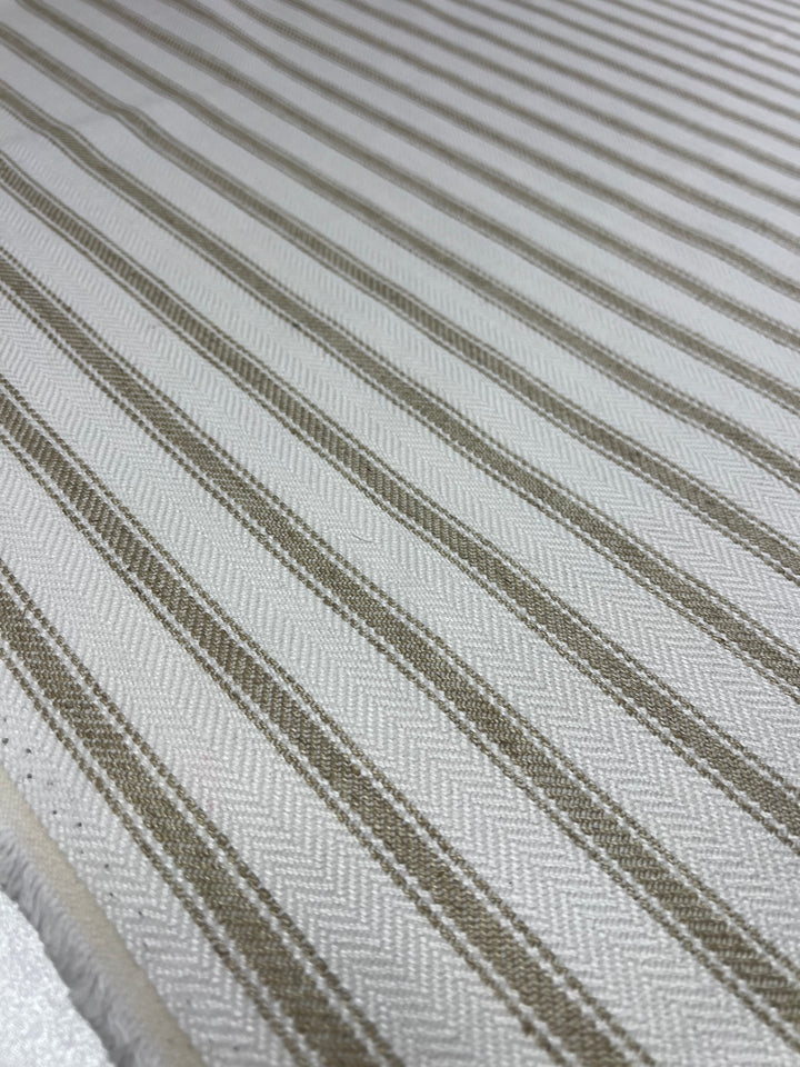 Close-up of a textured upholstery fabric featuring a herringbone pattern with alternating light and dark beige stripes running diagonally across the surface. The durable, stylish fabric appears soft and neatly woven, with a slight sheen under the light—perfect for an affordable range from Super Cheap Fabrics' Upholstery Herringbone - Salt & Sand - 145cm collection.
